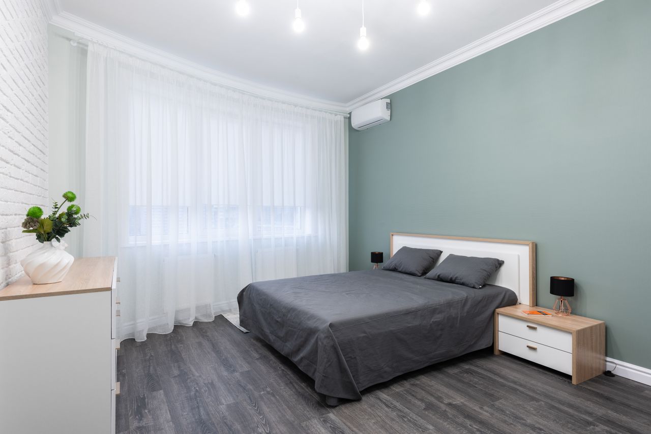 Contemporary bedroom with pillows and cover on bed against table with lamp and decorative plant on commode